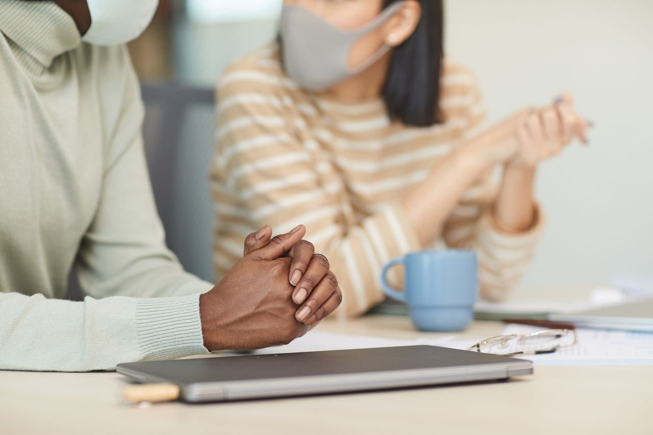 Two colleagues wearing face masks, sitting and discussing work at a desk in a modern office setting.