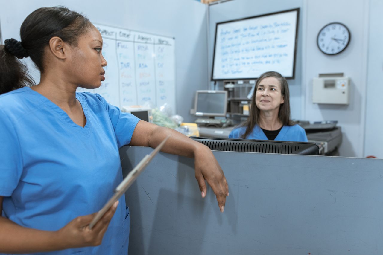 Nurses in scrub suits discussing medical tasks in a clinical environment.